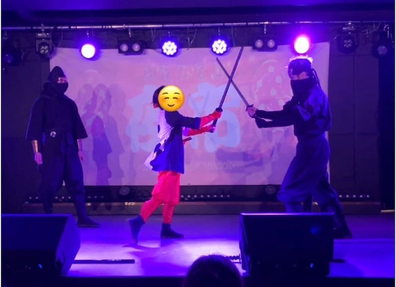 A stage performance with performers in costumes, a large screen in the background, and stage lighting. 1