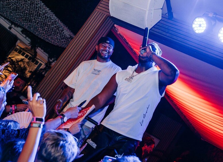 Two people in white t-shirts, one holding a microphone, and a crowd of people with raised hands. 3