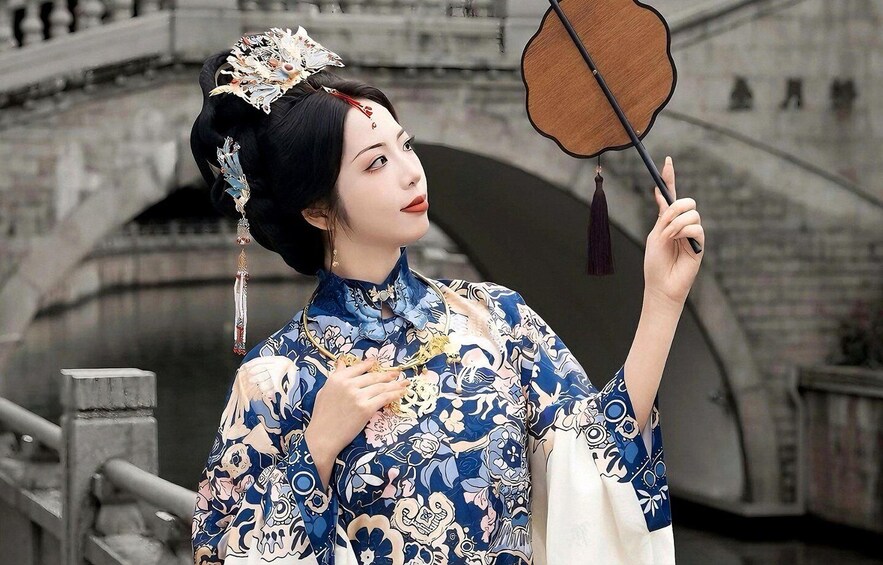 A person in traditional attire holding a fan, with a bridge and stone architecture in the background. 1