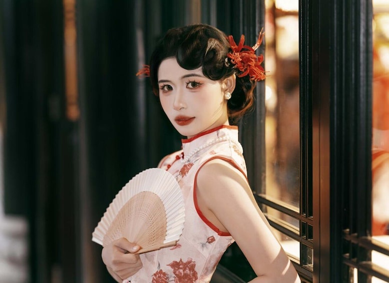 A person in traditional attire holding a fan, standing by a window with a view of buildings. 1