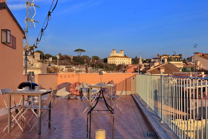A rooftop terrace with a table and chairs, overlooking a cityscape with historic buildings. 1