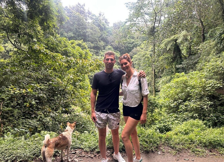 A man and woman standing in a forested area with a dog nearby. 3