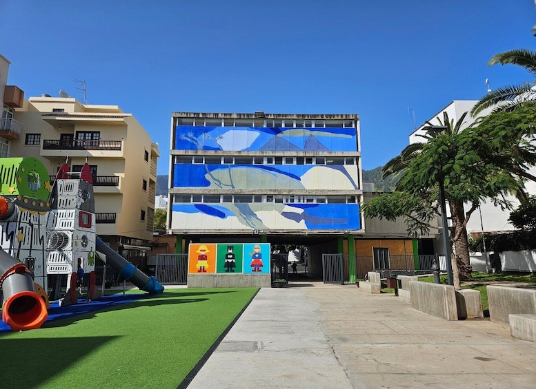 A playground with a slide and artificial turf, a multi-story building with colorful murals, and a palm tree. 2