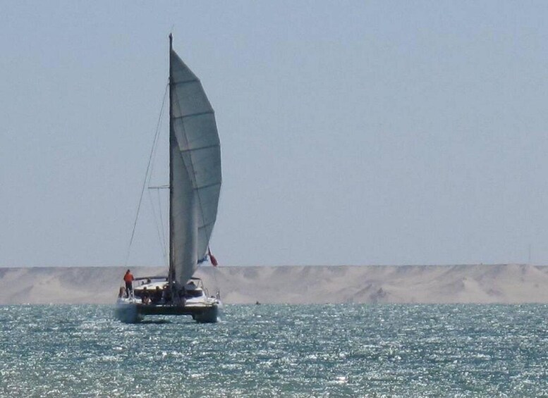 A sailboat with a crew on a calm sea with a sandy shore in the background. 2