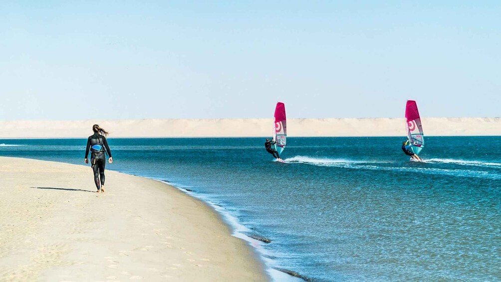 A beach with two windsurfers and a person walking along the shore. 4