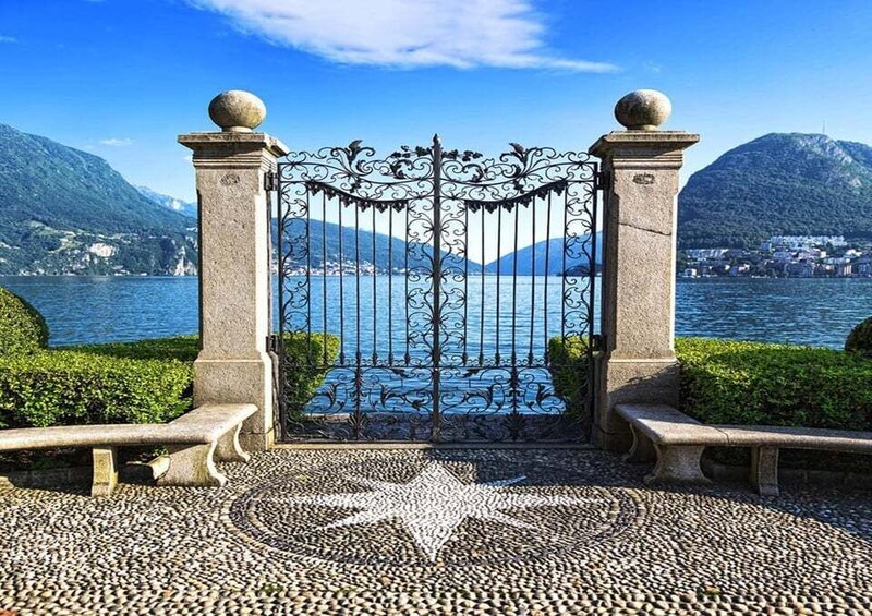 A stone gate with ornate metalwork, leading to a lake with mountains in the background. 5