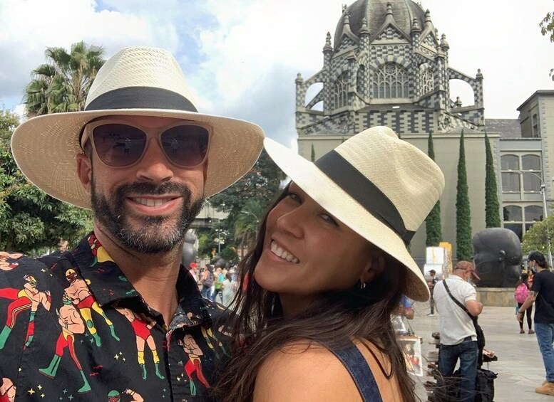A couple wearing hats, a man in a colorful shirt, and a woman in a black top, with a large, ornate building in the background. 1