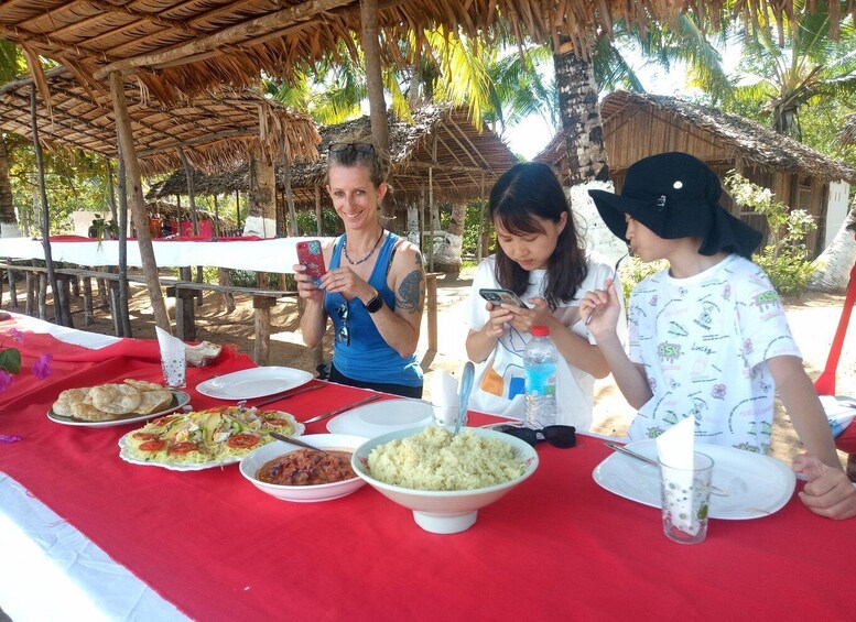 A group of people dining outdoors under a thatched roof, with a table set with various dishes and a tropical backdrop. 2
