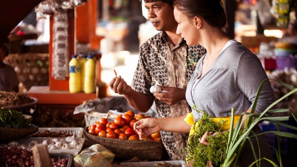 A person holding a garlic bulb and another person holding a red chili pepper at a market stall with various vegetables and spices. 1