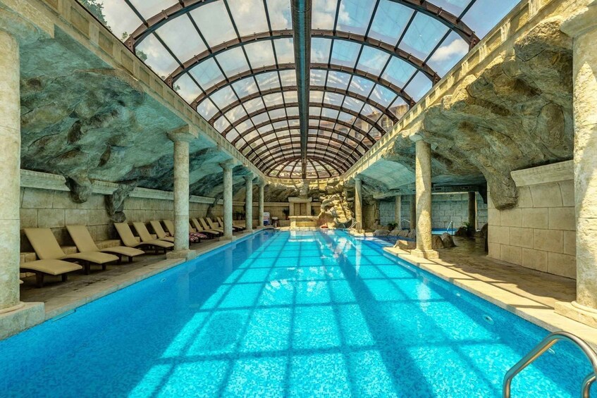 An indoor swimming pool with a glass ceiling, stone walls, and lounge chairs. 3