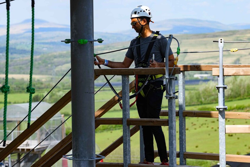 A person on a wooden platform with metal railings, wearing a helmet and harness, overlooking a green landscape with hills. 5