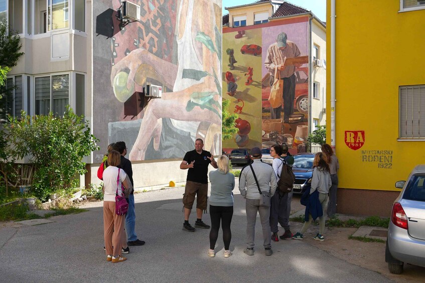 A mural on a building, a group of people standing in front of it, and a yellow wall with graffiti. 2