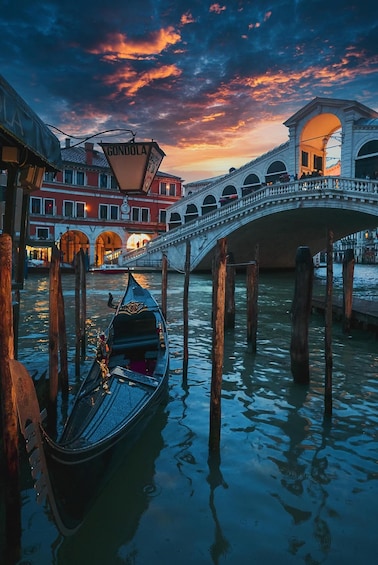 A gondola docked at a bridge in a canal with a sunset sky. 1