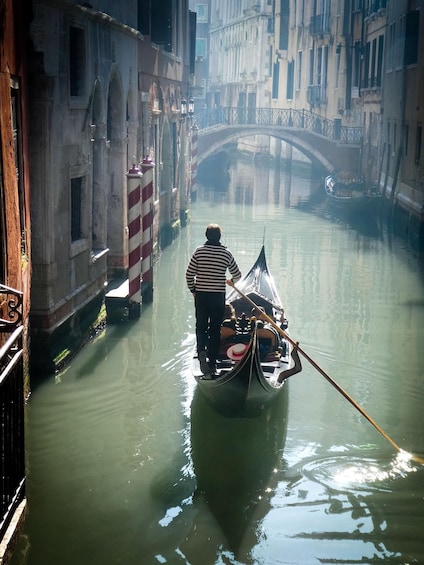 A gondolier in a narrow canal with buildings and a bridge in the background. 3