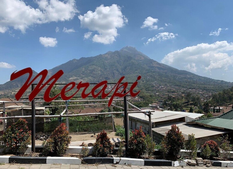 A sign with 'Merapi' written on it, a mountain in the background, and a town below. 4