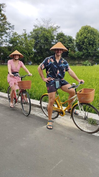 Two people on bicycles with baskets, wearing hats, and standing by a road with green fields and trees in the background. 5