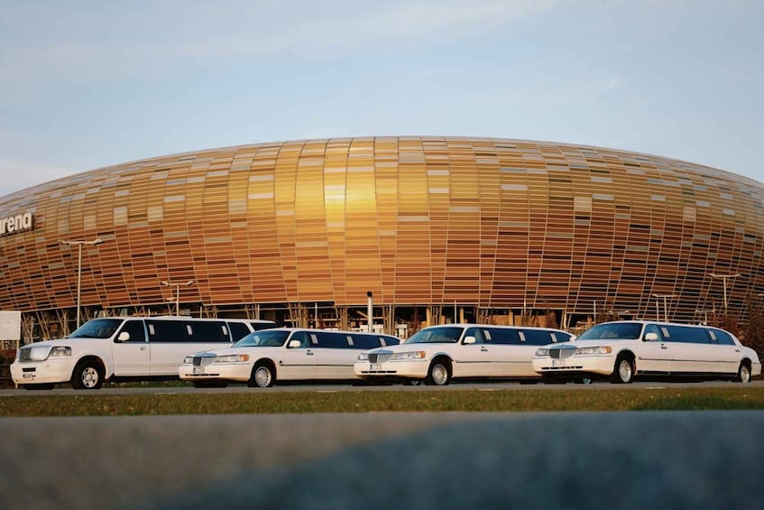 A large, modern stadium with a distinctive ribbed facade and a row of white limousines parked in front. 4