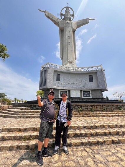 Two people standing in front of a large statue with outstretched arms, a building with inscriptions, and a stone staircase. 3