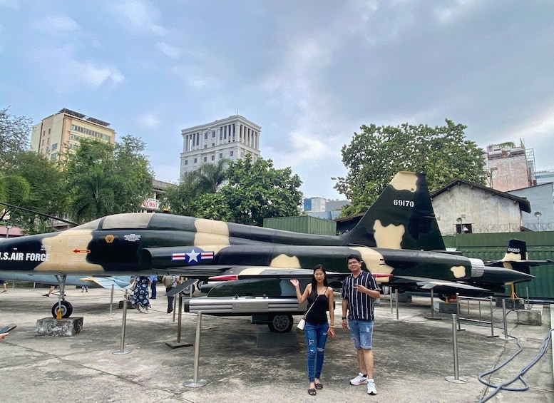 A military aircraft with camouflage paint and a star insignia, parked outdoors with a cityscape in the background. 1