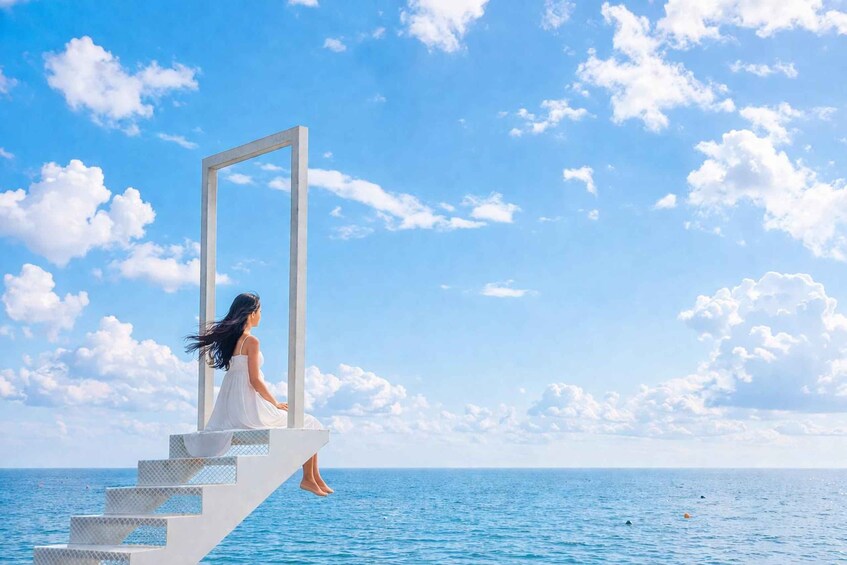 A woman in a white dress sitting on a staircase by the sea, looking out at the ocean. 1