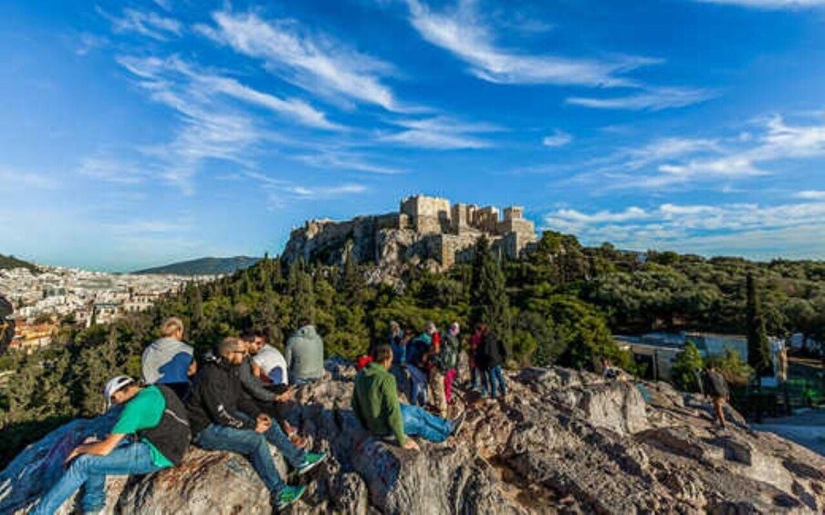 A group of people sitting and standing on a rocky hill overlooking a historic castle on a hilltop. 4