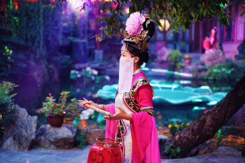 A person in traditional attire with a floral headdress, holding a red container, standing in a garden with rocks and a pond. 3