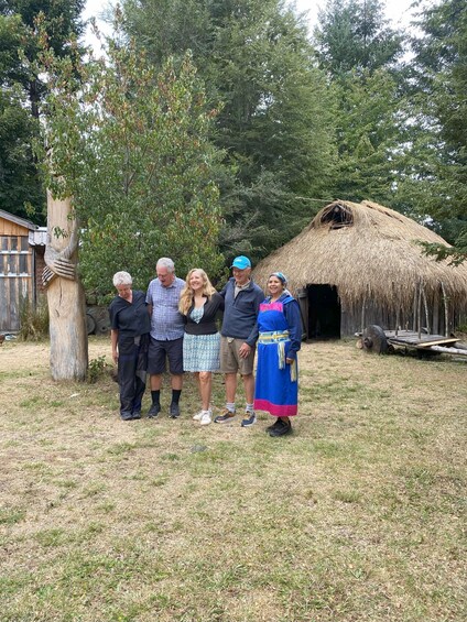 A group of people standing in front of a thatched-roof structure with a wooden wheel and a tree with carved bark. 4