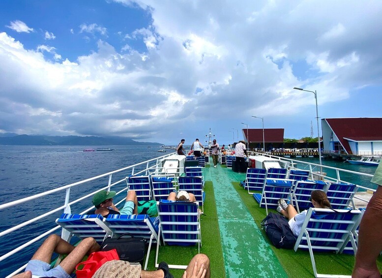 A ferry deck with passengers seated on blue chairs, a green pathway, and a body of water with boats in the distance. 4
