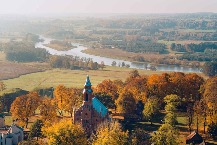 Een kerk met een groen dak en een toren, omgeven door herfstkleurige bomen en velden. 1