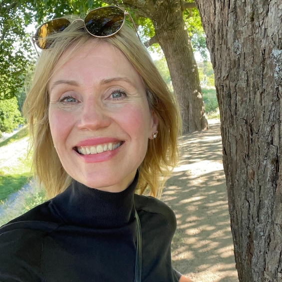 A person with blonde hair, wearing a black turtleneck, standing by a tree with a dirt path in the background. 1