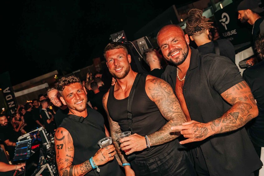 A group of people at an outdoor event, some with tattoos, and a DJ booth in the background. 3