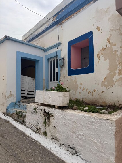 A weathered building with peeling paint, a blue door frame, a window with a pink frame, and a planter with flowers. 2