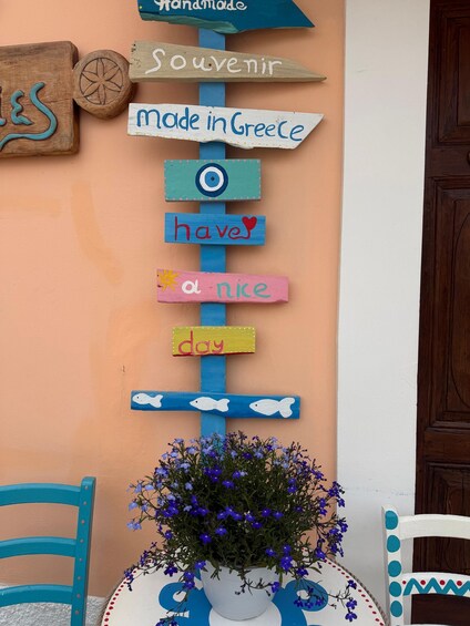 A vertical wooden sign with various phrases like 'Souvenir,' 'made in Greece,' and 'have a nice day,' against a peach wall. 3