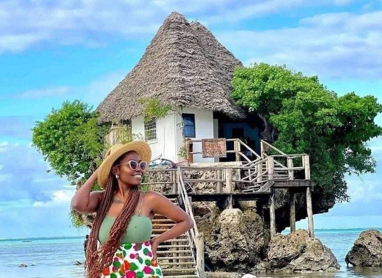 A woman in a tropical setting with a thatched-roof house on stilts over water. 1