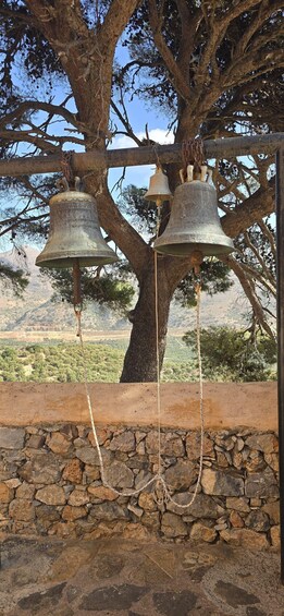 Three large bells hanging from a tree, with a stone wall and a mountainous landscape in the background. 2