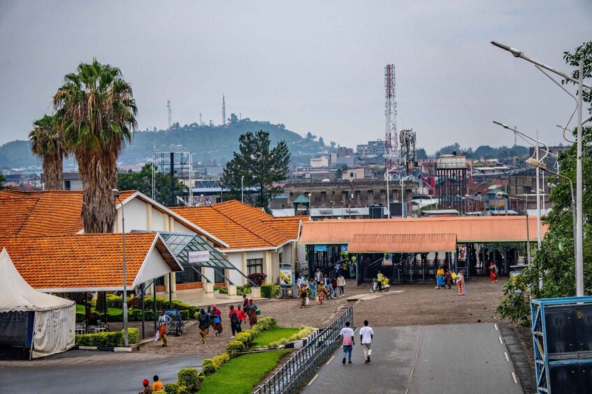 A town square with people walking, a palm tree, and buildings with orange roofs. 1
