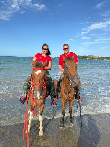 Two people riding horses on a beach with water and a distant shoreline. 4