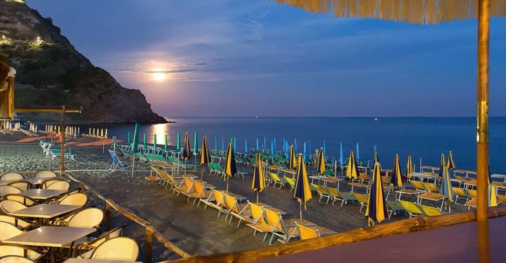 A beach with rows of sun loungers and umbrellas, a rocky cliff, and a calm sea under a twilight sky. 3