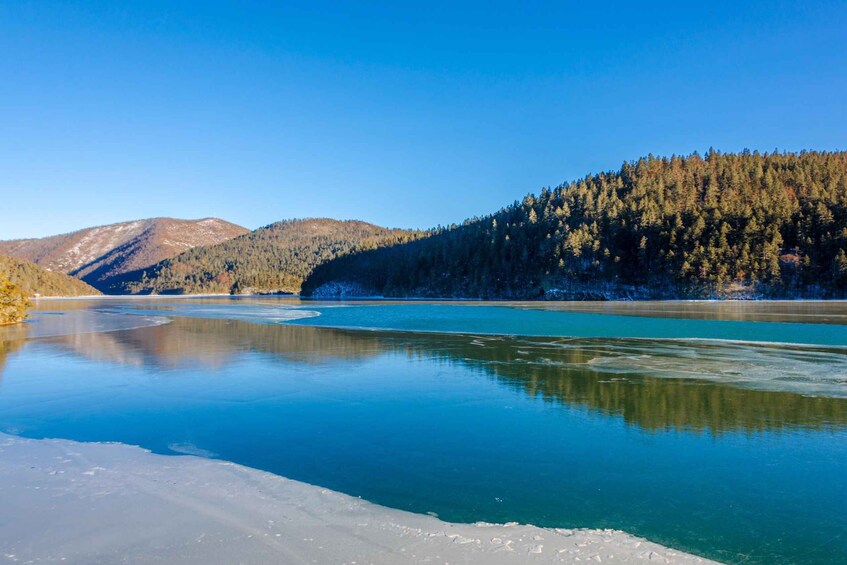 A frozen lake with clear blue water, surrounded by forested hills and mountains. 4