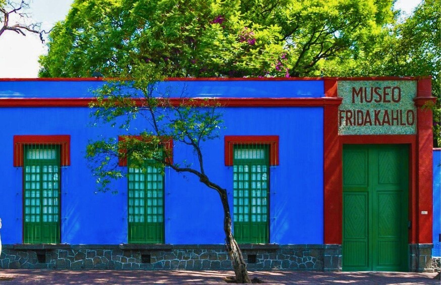 Sinisen julkisivun, vihreiden ovien ja ikkunoiden koristama rakennus, jonka yläpuolella on kyltti, jossa lukee: »MUSEO FRIDAKAHLO». 1