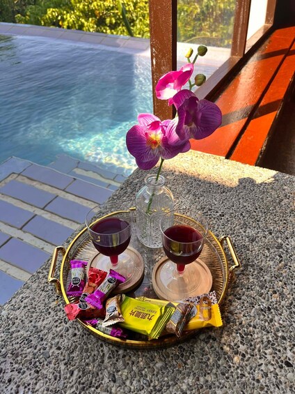 A tray with two glasses of red drink, a vase with purple flowers, and some snacks on a stone surface by a pool. 2