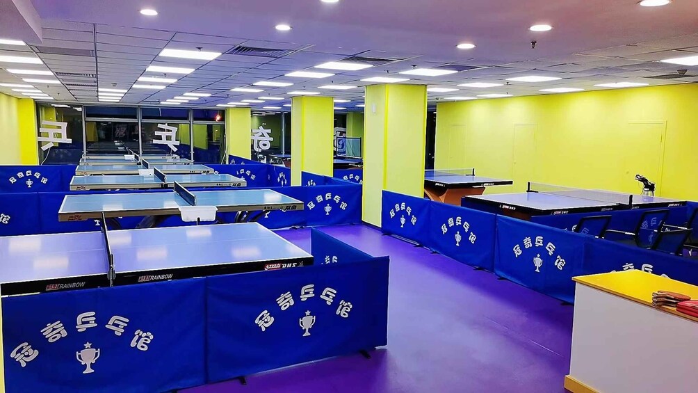 A brightly lit indoor table tennis facility with multiple tables and blue barriers. 5