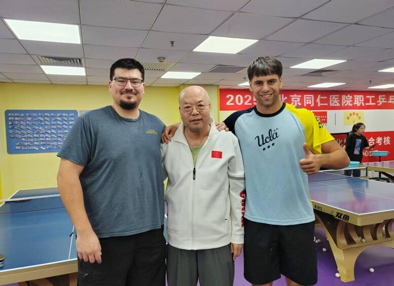 Three people standing in a room with a ping pong table, a red banner, and a bulletin board with papers. 3