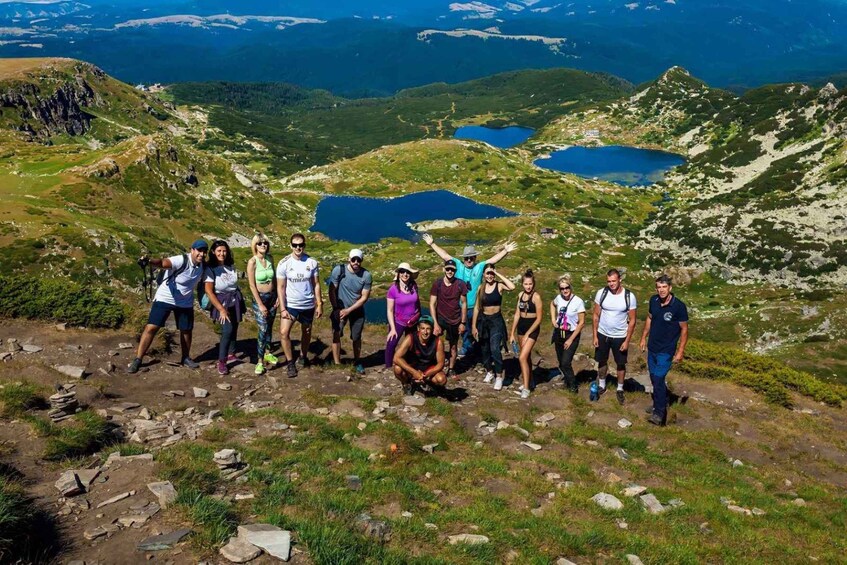 A group of people on a mountain trail with a lake and rocky terrain in the background. 1