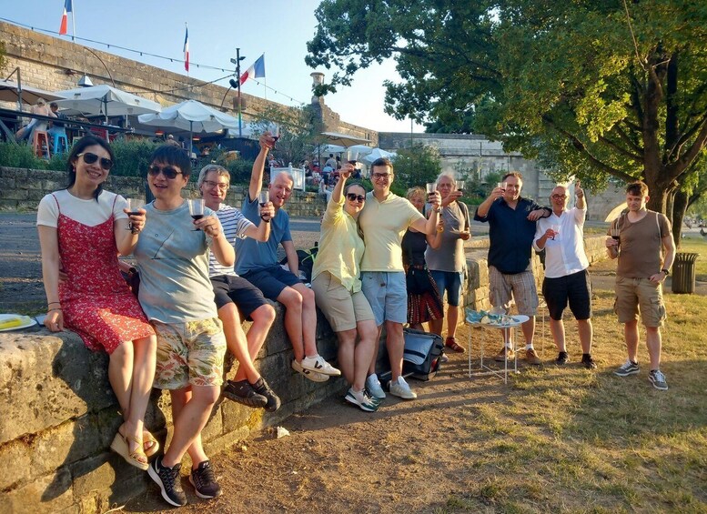 A group of people sitting and standing on a stone wall, holding drinks, with a flag and a building in the background. 1