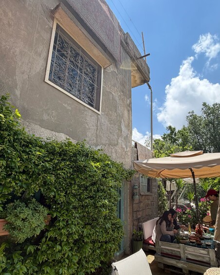 A rustic building with a decorative window, lush greenery, and a wooden table under a canopy. 2