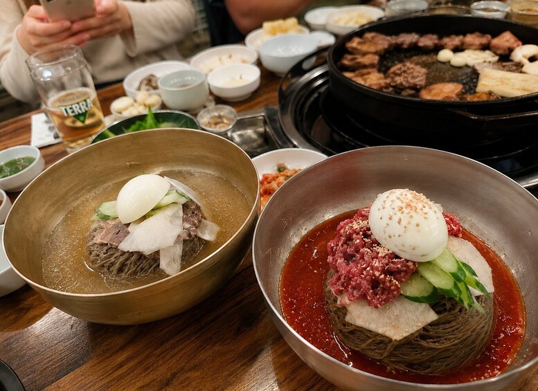 A table with two bowls of Korean dishes, one with a clear broth and another with a red sauce, and a black grill with meat in the background. 3