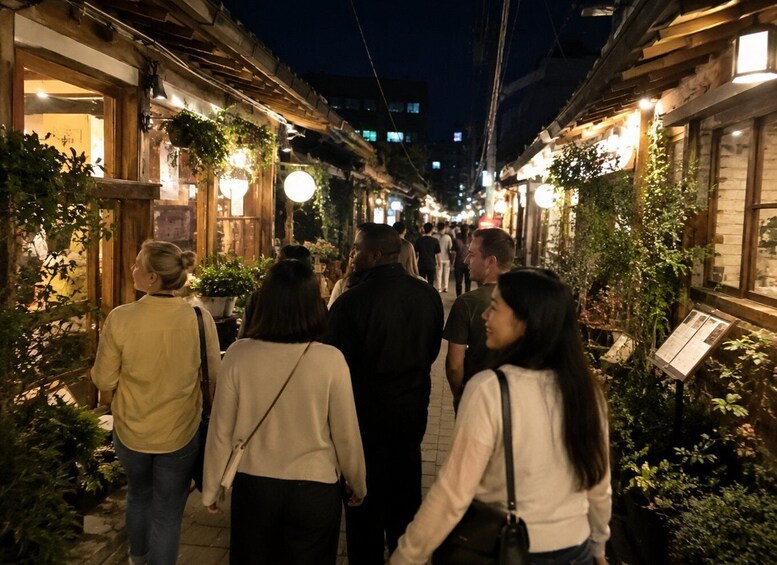 A nighttime street with people walking, illuminated by hanging lights and greenery. 2