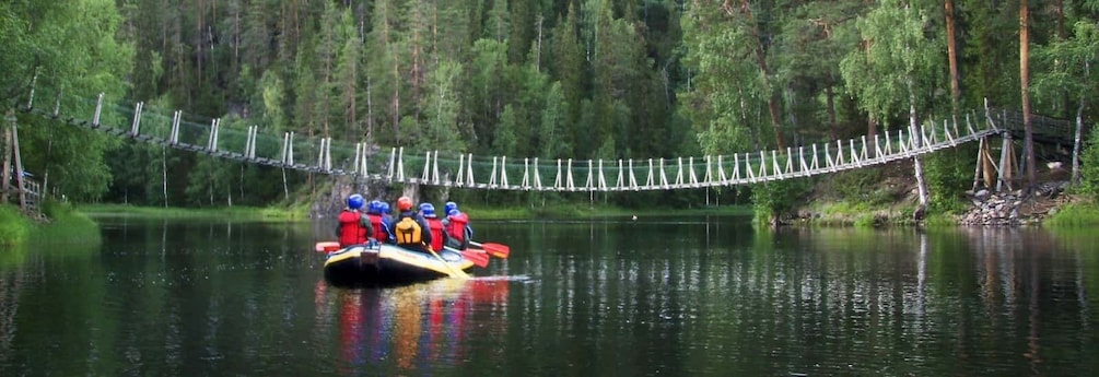 A group of people in a raft on a calm river, with a suspension bridge in the background. 1