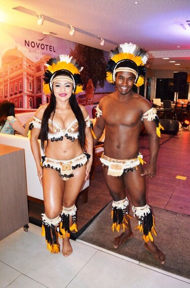 Two people in traditional attire with feathered headdresses, standing indoors with a hotel sign in the background. 2